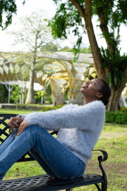 Parktaki bankta oturan genç bir kadın temiz havanın tadını çıkarıyor ve gökyüzüne bakıyor.