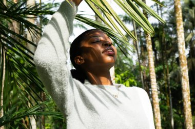 Palmiye yapraklarına dokunan, tropik bir ormanda güneş ışığının tadını çıkaran açık yeşil bir kazak giyen bir model.