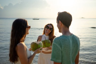 Altın saat boyunca elinde taze hindistan cevizi içkileriyle sahilde gülen üç arkadaş.