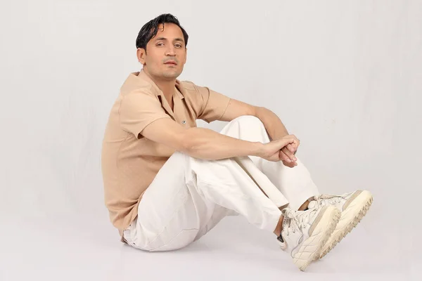 Beyaz gömlekli genç adam stüdyoda yerde oturuyor.