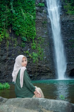 Asyalı Müslüman kadınlar, şelalenin ve yemyeşil ağaçların güzel zeminiyle bir kayanın üzerinde otururken zarif görünürler..