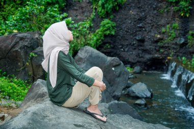 Asyalı Müslüman kadının nehir kenarındaki büyük bir kayanın üzerinde bacaklarını tutarken görüntüsü..