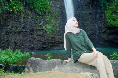 Asyalı Müslüman kadınlar, şelalenin ve yemyeşil ağaçların güzel zeminiyle bir kayanın üzerinde otururken zarif görünürler..