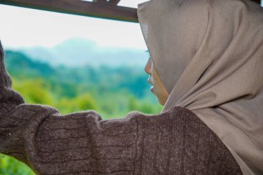 Asyalı Müslüman bir kadın yan profilde görünüyor, düşünceli bir şekilde tepe manzarasına bakıyor..