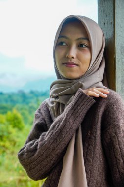 Asyalı Müslüman bir kadının sağ eliyle kameraya dönük pozu sol omzunu tepe manzarasının önünde tutuyor..