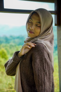Asyalı Müslüman bir kadının sağ eliyle kameraya dönük pozu sol omzunu tepe manzarasının önünde tutuyor..