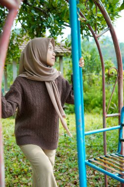 Asyalı Müslüman bir kadın, arkadaşça bir gülümsemeyle sallanan oyun alanında iki dik demir merdiven tutuyor..