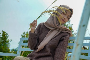 Asyalı Müslüman bir kadının, sakin bir ifadeyle ve elinde bir demet yabani otla güzel bir parkın ortasında, bir bankta otururken görüntüsü..