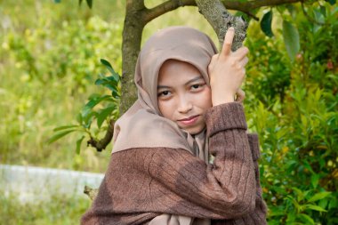 Asyalı Müslüman kadın, yapraklarla kaplı bir ağaç gövdesine yaslanarak kameraya nazikçe bakar..