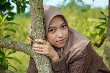 Asyalı Müslüman kadın, yapraklarla kaplı bir ağaç gövdesine yaslanarak kameraya nazikçe bakar..
