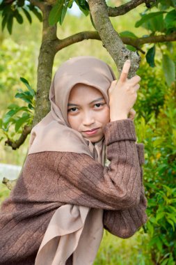 Asyalı Müslüman kadın, yapraklarla kaplı bir ağaç gövdesine yaslanarak kameraya nazikçe bakar..