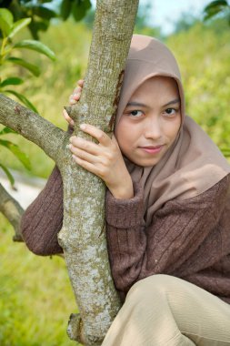 Asyalı Müslüman kadın, yapraklarla kaplı bir ağaç gövdesine yaslanarak kameraya nazikçe bakar..