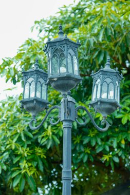 Viktorya dönemi tarzı işlenmiş demir çerçeveli bahçe lambası, bir adım çatı süsü tasarımı ve eğri cam paneller ile ön planda duruyor.