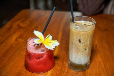 Tahta bir masada iki bardak soğuk içecek köpük serpiştirilmiş karpuz suyu ve frangipani çiçeği süslemesi ve siyah pipetli buzlu kahve, bir tropikal ve hakem.