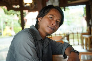 Uzun saçlı güneydoğu Asyalı bir adamın, sakin bir ifade ve hafif bir gülümsemeyle geleneksel ahşap mimari bir zemine karşı yan görünüşü..