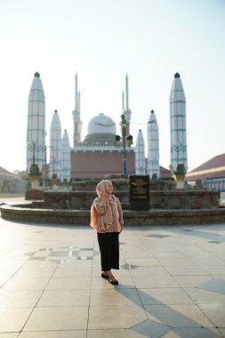 Kubbeli bir cami binası ve arka planda uzun minareler ile gülümseyen Asyalı Müslüman kadın görüntüsü.