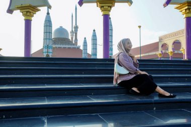 Asyalı Müslüman kadın caminin önünde mutlu bir gülümsemeyle oturuyor..