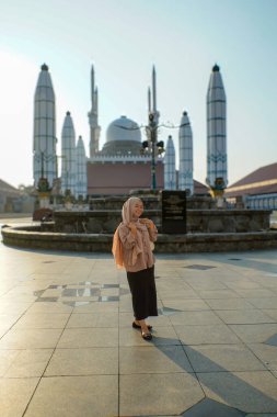 Kubbeli bir cami binası ve arka planda uzun minareler ile gülümseyen Asyalı Müslüman kadın görüntüsü.