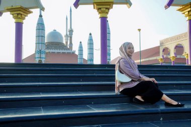 Asyalı Müslüman kadın caminin önünde mutlu bir gülümsemeyle oturuyor..