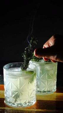 Cin tonik ve bardaklarda buzla yapılan nefis alkollü kokteyl geceleri dinlenmek ve yazın ferahlatıcı olmak için. Baharat ve aroma için yeşil limon. Barmen ve bar. Biberiye ateşle yanıyor.