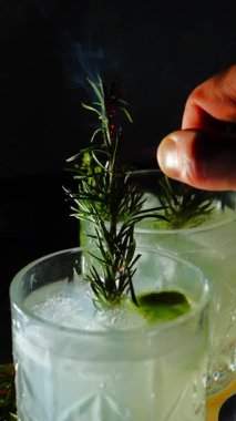 Cin tonik ve bardaklarda buzla yapılan nefis alkollü kokteyl geceleri dinlenmek ve yazın ferahlatıcı olmak için. Baharat ve aroma için yeşil limon. Barmen ve bar. Biberiye ateşle yanıyor.