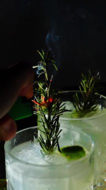 Cin tonik ve bardaklarda buzla yapılan nefis alkollü kokteyl geceleri dinlenmek ve yazın ferahlatıcı olmak için. Baharat ve aroma için yeşil limon. Barmen ve bar. Biberiye ateşle yanıyor.