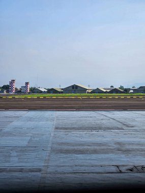 Bir uçak hangarının içinden açık hava sahasına dikey bir bakış açısı. Beton zemin hatları uzak hangarlara, kontrol kulelerine ve temiz mavi gökyüzüne götürür, havacılık altyapısını, teknik operasyonları ve havaalanına hazır olmayı temsil eder..