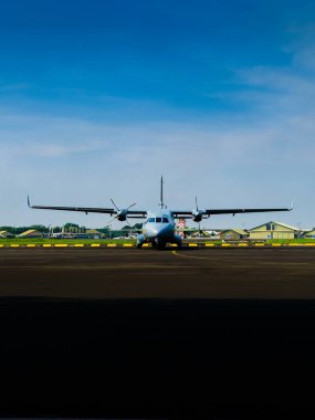 Pist merkezine yerleştirilmiş kargo pervane uçağının göz hizasında bir perspektifi. Yüksek kontrastlı gün ortası aydınlatması ön planda kısmi siluet oluşturur, hazırlık, havacılık gücü ve görev öncesi durgunluğu iletir.