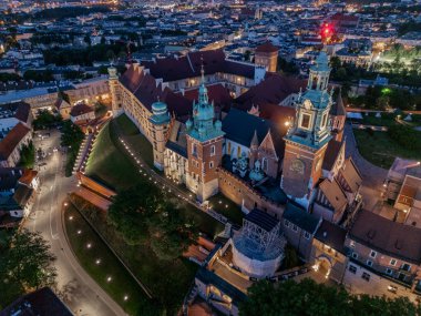 Krakow 'daki Wawel Kalesi' nin (Krakow) alacakaranlıkta çekilmiş insansız hava aracı görüntüleri, arka planda aydınlanmış duvarlar, Vistula Nehri ve şehrin ufuk çizgisini gösteriyor..