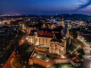 Krakow 'daki Wawel Kalesi' nin (Krakow) alacakaranlıkta çekilmiş insansız hava aracı görüntüleri, arka planda aydınlanmış duvarlar, Vistula Nehri ve şehrin ufuk çizgisini gösteriyor..