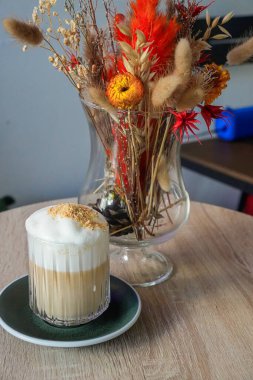 Bir vazo kurutulmuş çiçeğin yanındaki masada beyaz köpüklü latte.