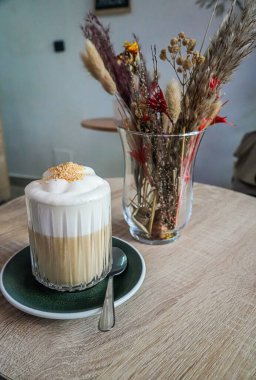 Beyaz köpüklü latte yakın bir yerde vazo dolusu kurumuş çiçekle bir masada..