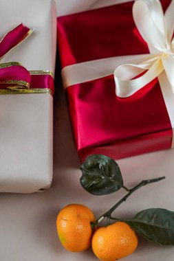 Tarafsız bir arkaplanda yan yana yerleştirilmiş, biri kırmızı diğeri bej olmak üzere iki hediye kutusunun yakın görüntüsü. Yanlarında yeni bir mandalina yatar ve Noel, Yeni Yıl veya doğum günü temalarına uygun bayram kompozisyonu oluştururlar..
