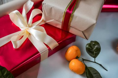 Üzerinde bej renkli bir hediye kutusu olan kırmızı hediye kutusunun yakından görüntüsü, her ikisi de kurdelelerle süslenmiş. Zirvede yeni bir mandalina, Noel, yeni yıl, tatil ve doğum günü temalarına uygun sıcak ve şenlikli bir kompozisyon oluşturur.