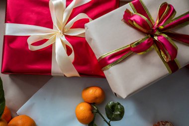 Üstünde bej renkli bir hediye kutusu olan, kurdelelerle süslenmiş kırmızı bir hediye kutusunun yakın görüntüsü. Zirvede yeni bir mandalina, Noel, Yeni Yıl, tatiller ve doğum günü temaları için minimal ve şenlikli bir kompozisyon oluşturuyor..