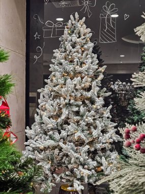 Suni karla kaplı dekoratif Noel ağacı hediye resimleriyle dekore edilmiş bir mağazanın camına dayanıyor. Bayram görselleri, mevsimlik dekorasyonlar, Noel gösterileri ve kış temalı projeler için ideal bir ortam..