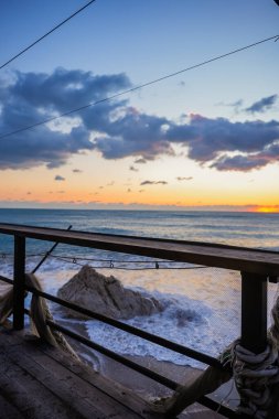 Gün batımında dalgalı bir denize bakan terastan dikey manzara. Sıcak turuncu ışık, hareket eden su ve karamsar kıyı atmosferi..