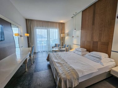 İçinde bir yatak, iki mavi ve beyaz sandalye, bir masa, yer lambası ve panoramik cam pencerelerin yanında uzun bir makyaj masası bulunan geniş bir otel veya ev manzarası. İç mekan, mimari ve yaşam tarzı fotoğrafçılığı için mükemmel..