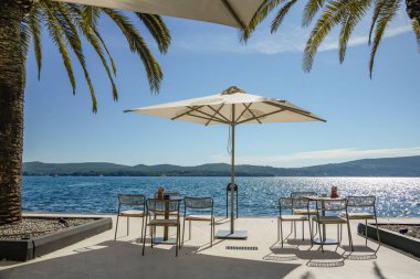 Rıhtımdaki açık kafe terasında ön planda palmiye yapraklarıyla çerçevelenmiş büyük bir şemsiye altında bir masa ve sandalyelerle. Güneşli Akdeniz sahnesi seyahat, yaşam tarzı, tatil ve eğlence içeriği için idealdir.