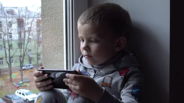 Băiat de patru ani cu smartphone pe pervazul ferestrei. Vederea parcului de toamnă și a spațiului stradal pentru text. Pericolul internetului Fotografie de stoc