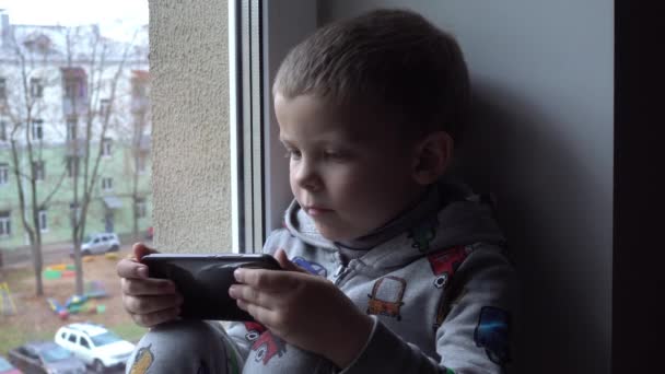 Bambino di quattro anni con smartphone sul davanzale della finestra. Vista del parco autunnale e spazio stradale per il testo. Pericolo di internet — Video Stock