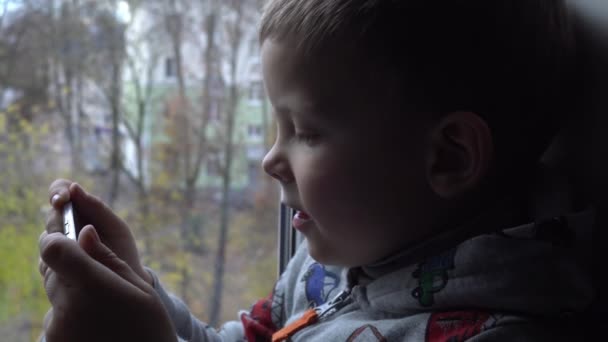 Čtyřletý kluk s chytrým telefonem na parapetu. Pohled na podzimní park a pouliční prostor pro text. Nebezpečí internetu — Stock video