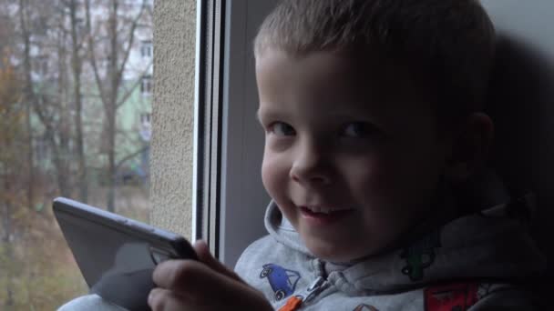Fyra år gammal pojke med smartphone på fönsterbrädan. Utsikt över höstparken och gatuutrymme för text. Fara för internet — Stockvideo