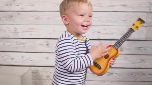 Niño feliz de dos años toca su guitarra o ukelele y canta canciones, sentado en la habitación de madera. — Vídeo de stock