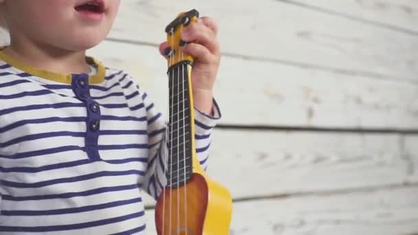 Szczęśliwy dwuletni chłopiec gra na gitarze lub ukulele i śpiewa piosenki, siedzi w drewnianym pokoju. — Wideo stockowe