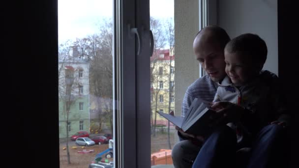父と息子は本を読んで写真から物語を作っています。壁と窓が背景にある — ストック動画