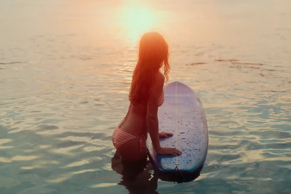 Surfer fată în bikini în picioare în apă, cu o placă de surf la apus de soare cu reflecție la soare — Fotografie, imagine de stoc
