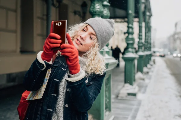 Joven turista rubia rizada con ropa de abrigo y guantes rojos fotografías o tomar una selfie en la calle de la ciudad, invierno, nieve —  Fotos de Stock