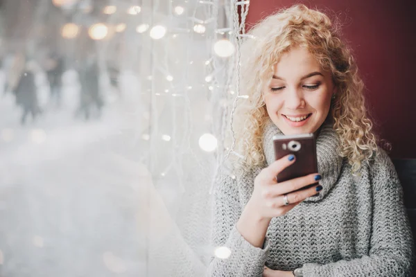 Joven rubia rizada hembra en suéter caliente usando un teléfono inteligente y sonriendo en la cafetería, ciudad de invierno fuera de la ventana —  Fotos de Stock