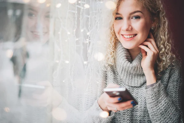 Joven rubia rizada hembra en suéter caliente usando un teléfono inteligente y sonriendo en la cafetería, mirando a la cámara, ciudad de invierno fuera de la ventana —  Fotos de Stock